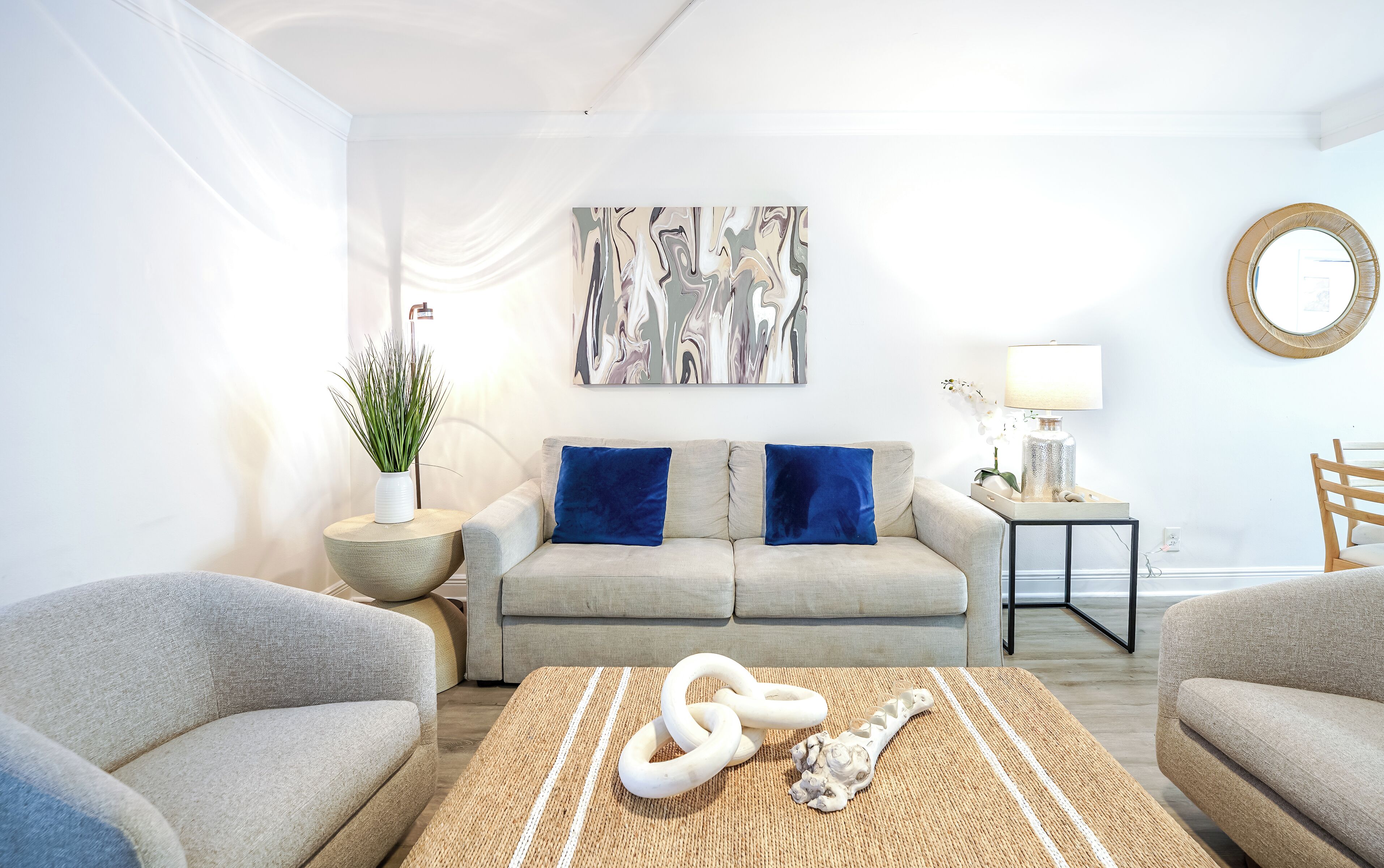 Uma sala de estar moderna com um sofá, duas poltronas, uma mesa de centro com objetos decorativos, uma mesinha lateral com abajur e um quadro abstrato pendurado na parede.