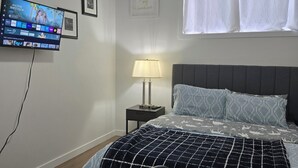 Un dormitorio con cama, escritorio, silla, televisor montado en la pared, lámpara de noche y ventana con cortinas.