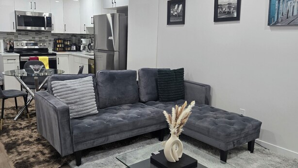 Una sala de estar moderna con un sofá seccional gris, una mesa de centro de vidrio y una cocina al fondo.