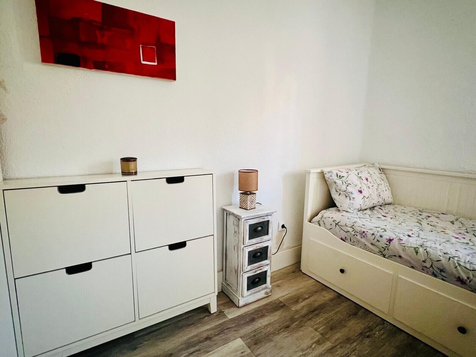 A bedroom with a bed, a white dresser with drawers, a small white cabinet with drawers, and a red abstract painting on the wall.