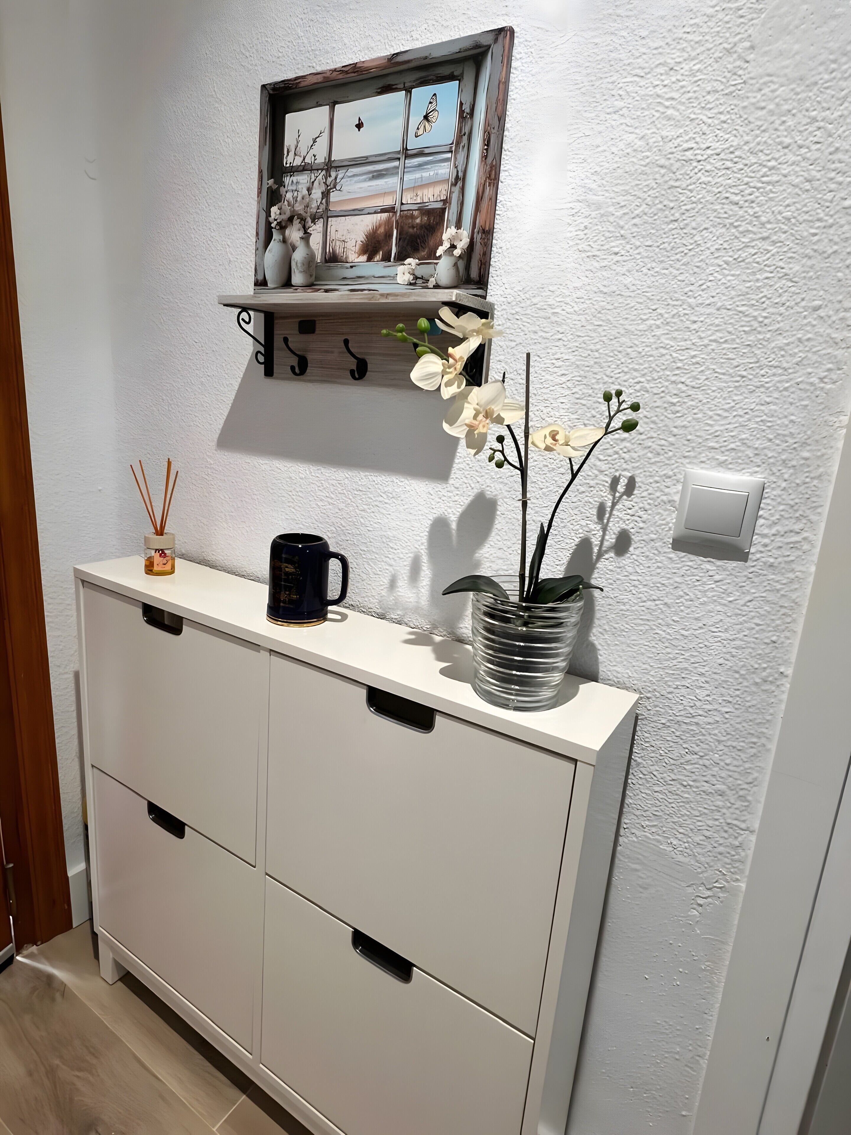 Een witte kast met lades, een zwarte mok, een glazen vaas met witte bloemen, een rietverspreider en een klein plankje met een fotolijst.
