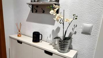 Een witte kast met lades, een zwarte mok, een glazen vaas met witte bloemen, een rietverspreider en een klein plankje met een fotolijst.