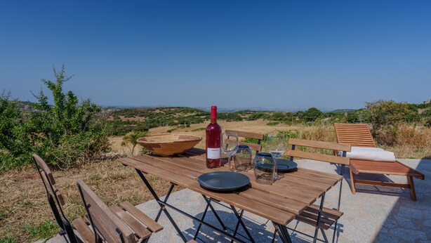 Ein Essbereich im Freien mit Tisch, Stühlen, einer Weinflasche, Gläsern und Blick auf die Landschaft.