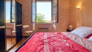 Un dormitorio con una cama grande, una ventana con cortinas, una silla y una mesita de noche con una lámpara.