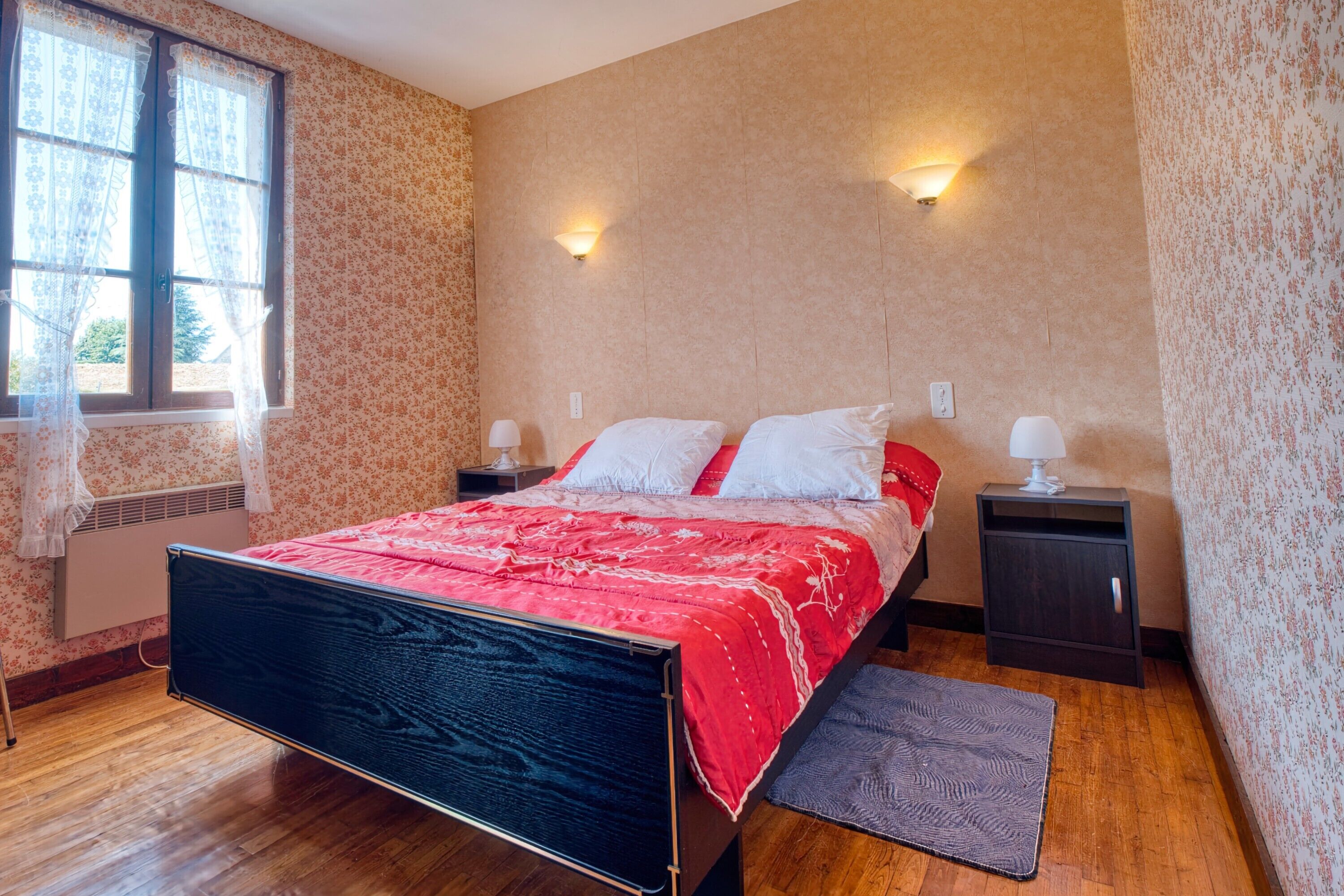 A bedroom with a bed, nightstands, a window with curtains, and a wall-mounted light fixture.