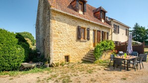 Una casa de piedra con techo de tejas, un pequeño porche y un espacio de comedor con mobiliario exterior.