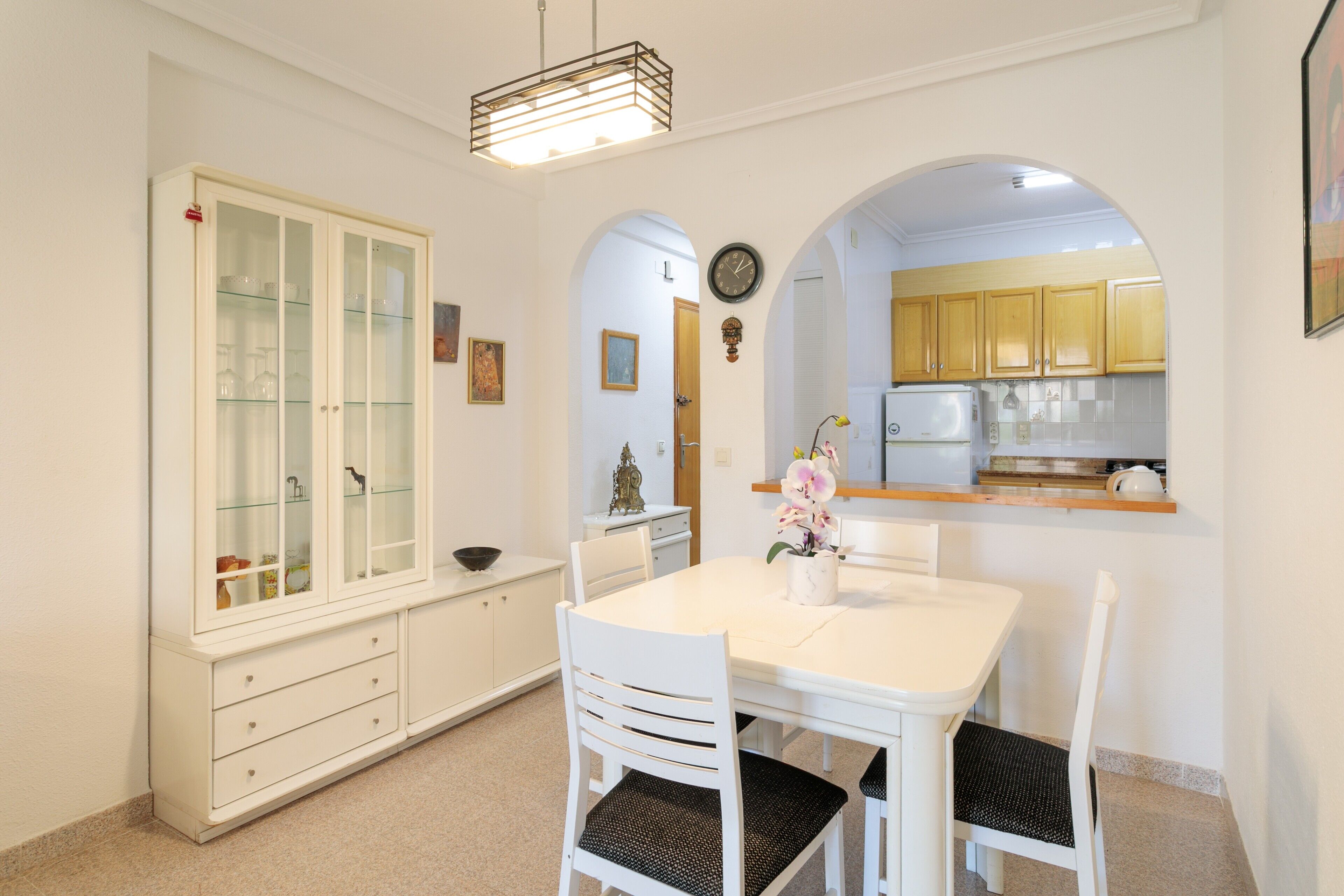 Un coin repas avec une table blanche, des chaises et un buffet aux portes vitrées, et une cuisine visible à travers une arche.