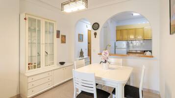 Un coin repas avec une table blanche, des chaises et un buffet aux portes vitrées, et une cuisine visible à travers une arche.
