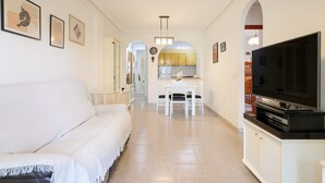 Una sala de estar moderna con un sofá, un comedor con mesa y sillas, una cocina al fondo y un televisor sobre un mueble.