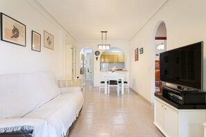 A modern living room with a sofa, a dining area with a table and chairs, a kitchen in the background, and a TV on a stand.