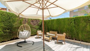 Un patio exterior con una sombrilla blanca, mobiliario de mimbre y un seto bien cuidado.