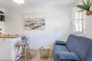 Moderni olohuone, jossa on harmaa sohva, pieni pyöreä pöytä, jonka päällä on kirja, tuoli, seinällä oleva maalaus, jossa on kuvattuna veneitä, ja kasvi ikkunalaudalla.