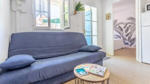 Ein modernes Wohnzimmer mit einem grauen Sofa, einem runden Couchtisch mit Zeitschriften, einem Fenster mit Vorhängen und einer Tür zu einem anderen Raum.