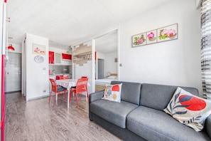 A modern living room with a grey sofa, red chairs, a dining table, and a kitchen in the background.