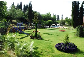A playground with slides and swings, a grassy area with flowers, and a building in the background.
