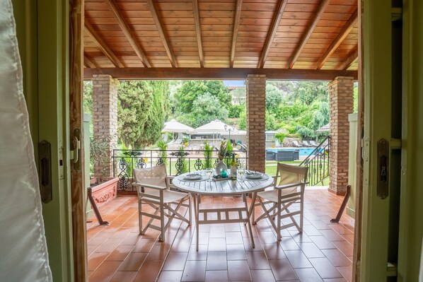 An outdoor patio with a wooden ceiling, a round table with chairs, and a view of a swimming pool and greenery.