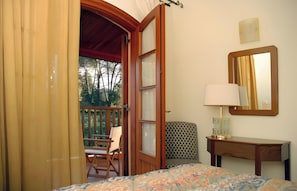 A bedroom with a bed, a bedside table with a lamp, a chair, a patterned sofa, a wooden door, and a balcony with a chair.