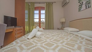 A hotel room with a bed, a desk with a television, a window with curtains, a wall-mounted air conditioner, and a painting.