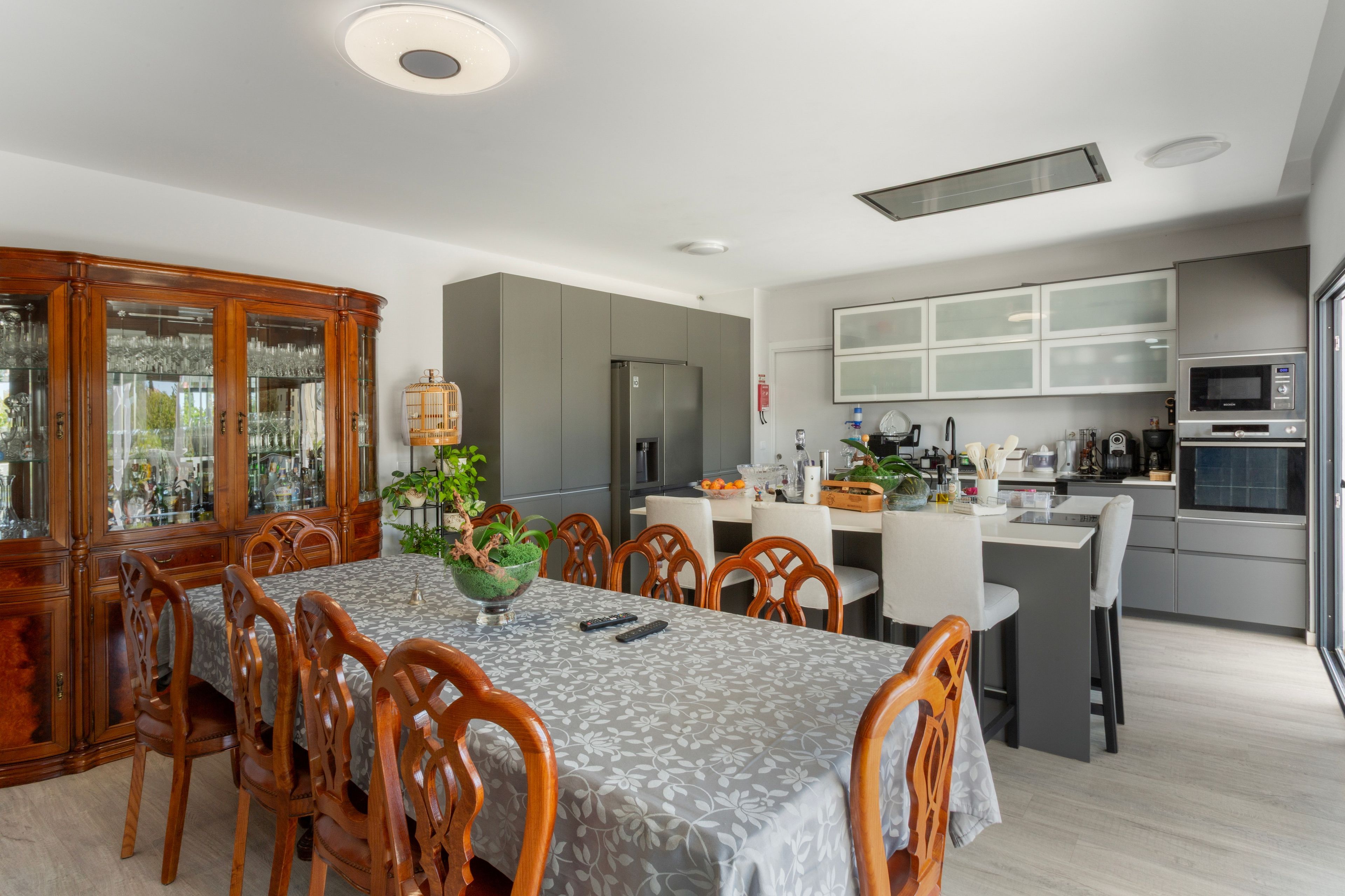 A modern kitchen with a built-in oven and microwave, a dining area with wooden chairs and a table, and a large cabinet with glass doors.
