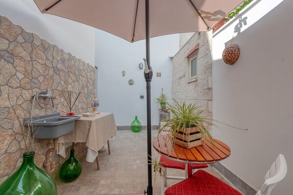 Un petit patio extérieur avec un mur en pierre, un évier, une table avec une chaise rouge et une plante en pot.