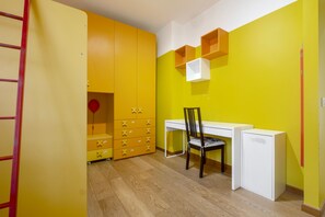 A room with a desk, chair, wardrobe, and a small white cabinet.