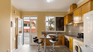A compact kitchen with a washing machine, two bar stools, a dining table, and a view through a glass door. - Apartment 'Casa Viva' with Sea View, Shared Terrace and Wi-Fi (Machico)