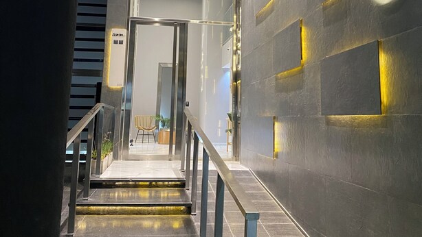 Una escalera moderna con barandillas de metal y peldaños iluminados, que conduce a un área de recepción con una pared oscura y lámparas con formas geométricas.