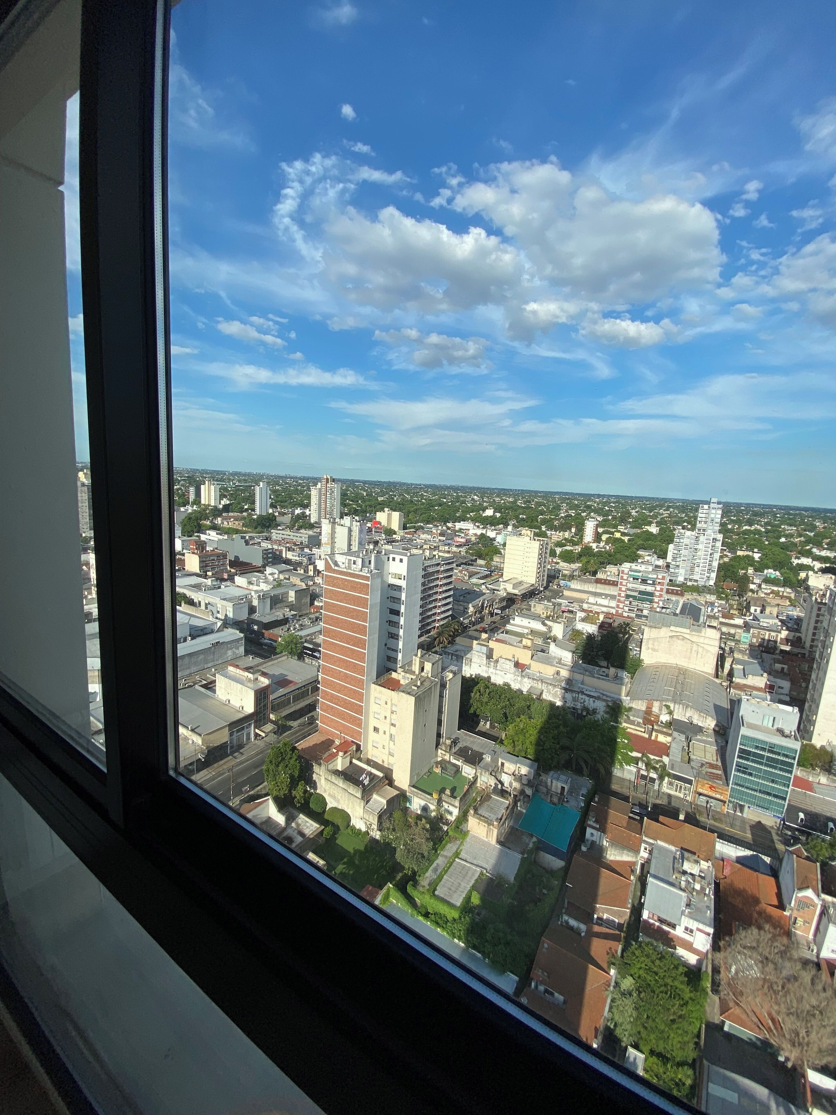 Une vue sur un paysage urbain, prise depuis une fenêtre, qui présente un mélange d’immeubles de grande hauteur et de bâtiments plus petits, avec un ciel dégagé et quelques nuages épars.