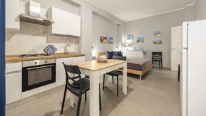 Una cocina compacta con estufa a gas, horno y gabinetes blancos. Cuenta con un comedor con mesa y sillas, y una cama con cabecera y almohadas.