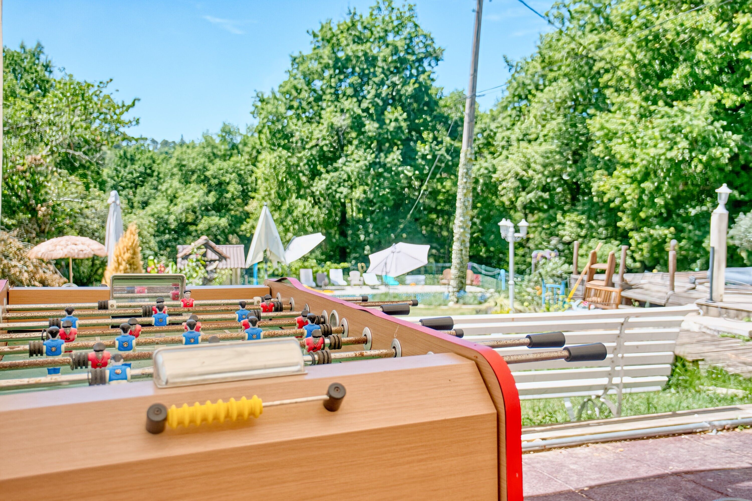 Une table de baby-foot en bois, avec un bord rouge, entourée d’un espace de détente extérieur et de verdure.