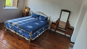 A bedroom with a bed, wooden floor, a window, a bedside table, and a mirror. - The House in the Green (Lortallo)