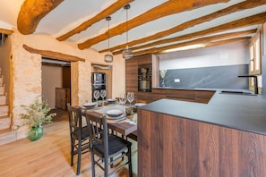 A modern kitchen with wooden beams, a dining area with a table set for two, and a stone wall. - Casa Rural El Ahora: Tradition, Art, and Design (Berge)