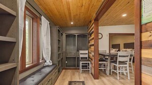 A cozy cabin interior with a wooden ceiling, a dining area with a table and chairs, a bench with storage, and a patterned rug on the floor. - Residential Area Ski Chalet by Creek - Sleeps 6 (Girdwood)