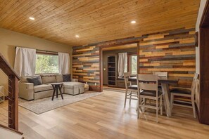 A spacious living area with a wooden ceiling, a sofa set, a dining table with chairs, and a staircase leading to another level. - Residential Area Ski Chalet by Creek - Sleeps 6 (Girdwood)