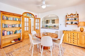 Uma sala de jantar com uma mesa redonda de madeira, cadeiras brancas, uma estante e uma cozinha visível através de um arco.