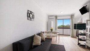 Una sala de estar moderna con un sofá negro, un comedor con mesa y sillas, una zona de cocina con mostrador y electrodomésticos, y un ventanal con vistas.