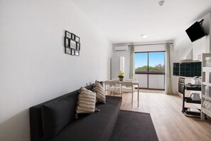 Una sala de estar moderna con un sofá negro, un comedor con mesa y sillas, una zona de cocina con mostrador y electrodomésticos, y un ventanal con vistas.