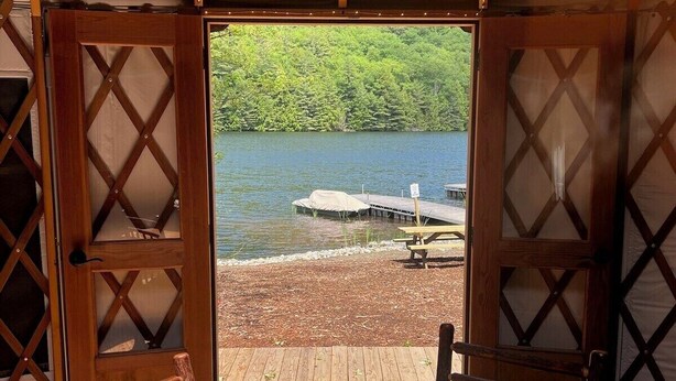 A wooden table with chairs and a vase of flowers, a wooden deck, and a view of a lake with a dock and greenery. - Shoreline Yurt @ Daggett Lake (Warrensburg)