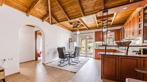 A spacious kitchen with wooden cabinets and a large island, a dining area with a table and chairs, and a view of the outdoors through a glass door. - Hillside Apartment Gjirokaster by PikHost (Gjirokastër)