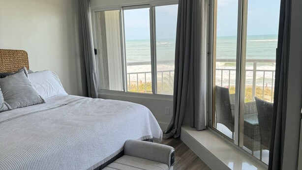 A bedroom with a large bed, a bench, a ceiling fan, and a view of the beach through large windows. - Beachfront 2BR/2BA Condo on Hutchinson Island w/ Private Garage (Fort Pierce)