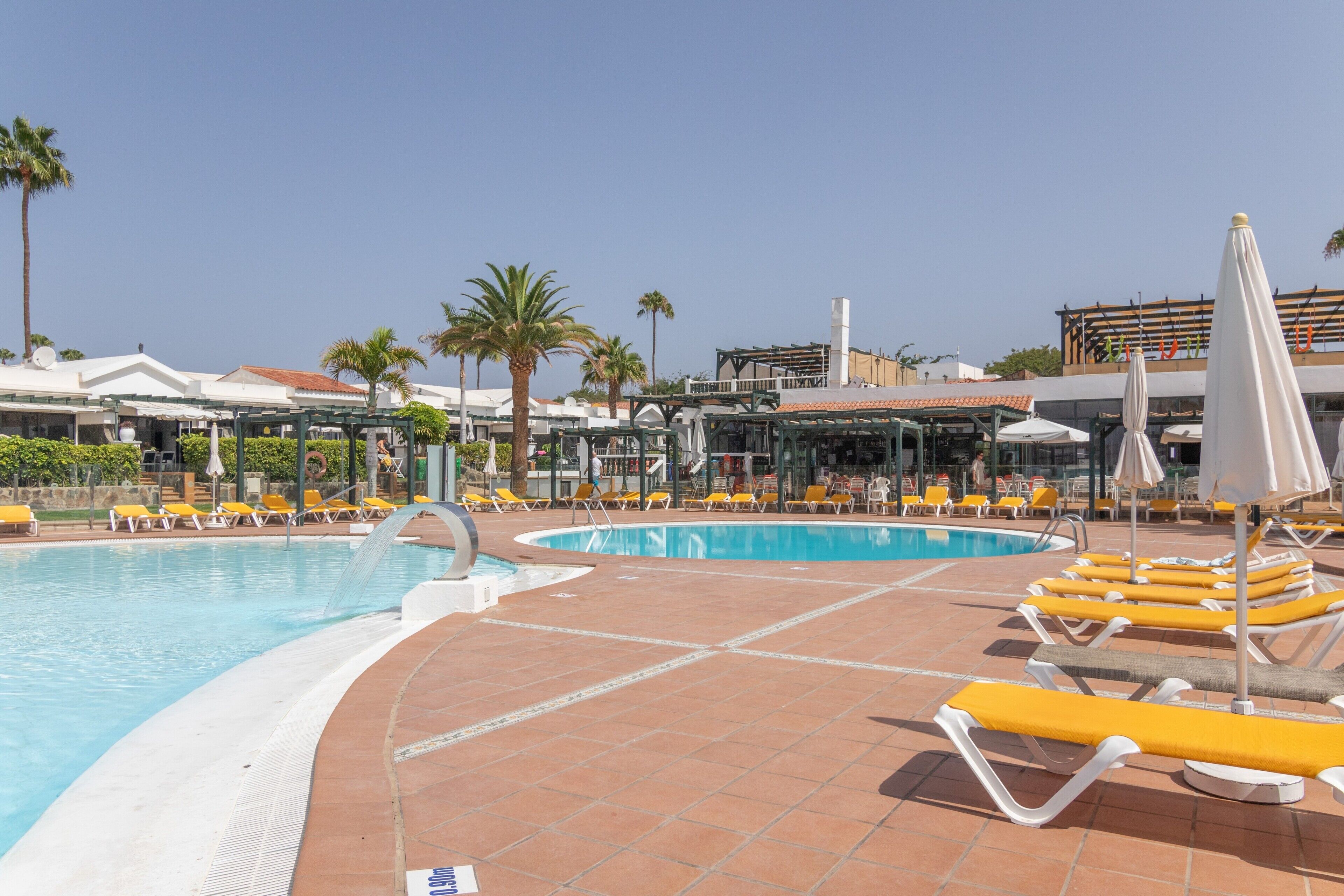 A swimming pool with sun loungers and umbrellas, surrounded by palm trees and buildings.