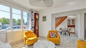 A modern living room with two yellow armchairs, a round glass table, a large window with a view of parked cars, a wooden staircase, and a dining area. - Holiday Home 'Maison Ker Hélène' with Private Terrace, Wi-Fi and Air Conditioning (Guérande)