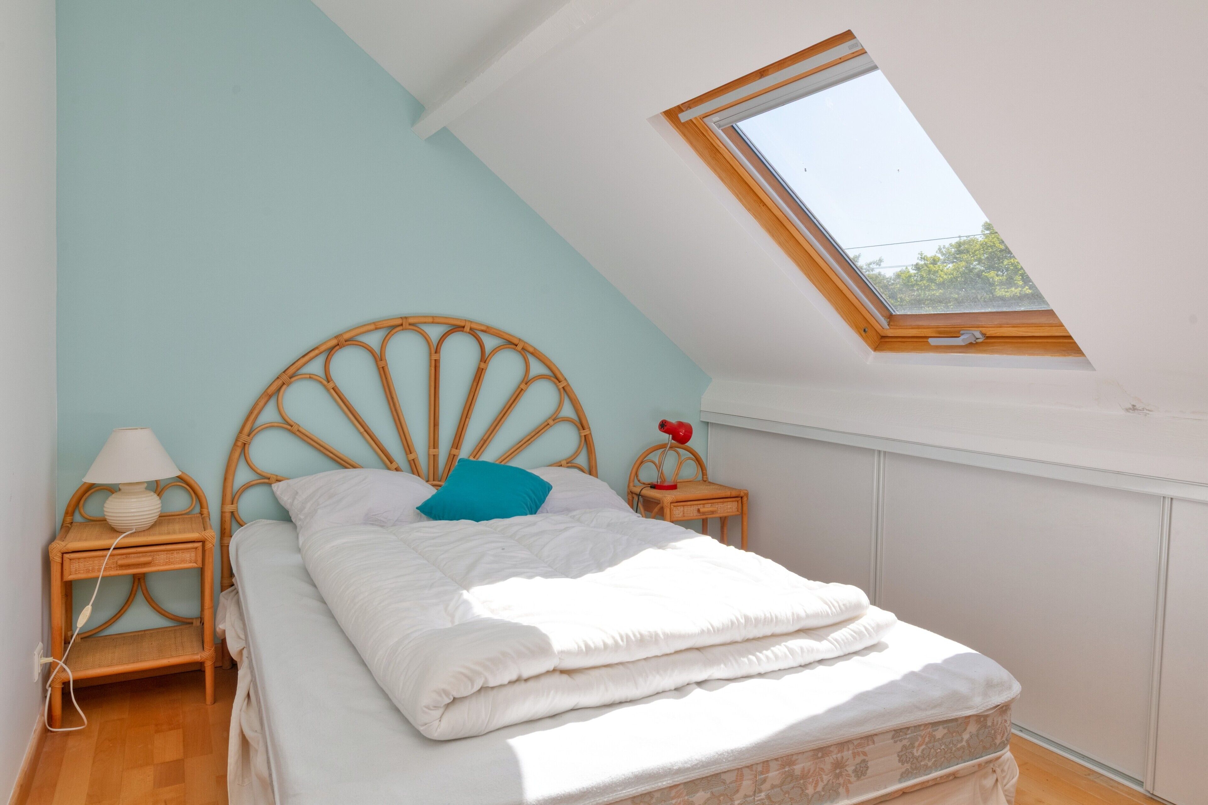 A bedroom with a skylight, a bed with white bedding, a wooden headboard, a nightstand with a lamp, and a small table with a red object.