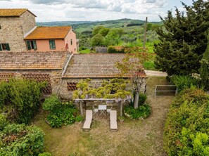 Un edificio rustico in pietra con tetto in tegole, circondato da vegetazione lussureggiante e una vista panoramica su dolci colline.