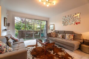 A spacious living room with a large sliding glass door leading to a backyard, a modern sofa set, a glass coffee table, and a decorative wall piece. - Unique Apartment with Balcony, Wi-Fi, and Air Conditioning (Hyères)