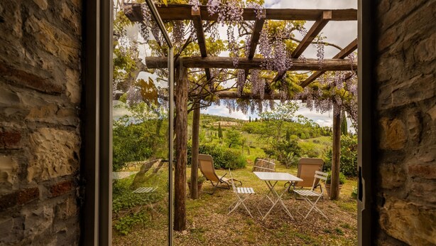 A stone-framed window opening to a garden with a pergola, chairs, and tables.