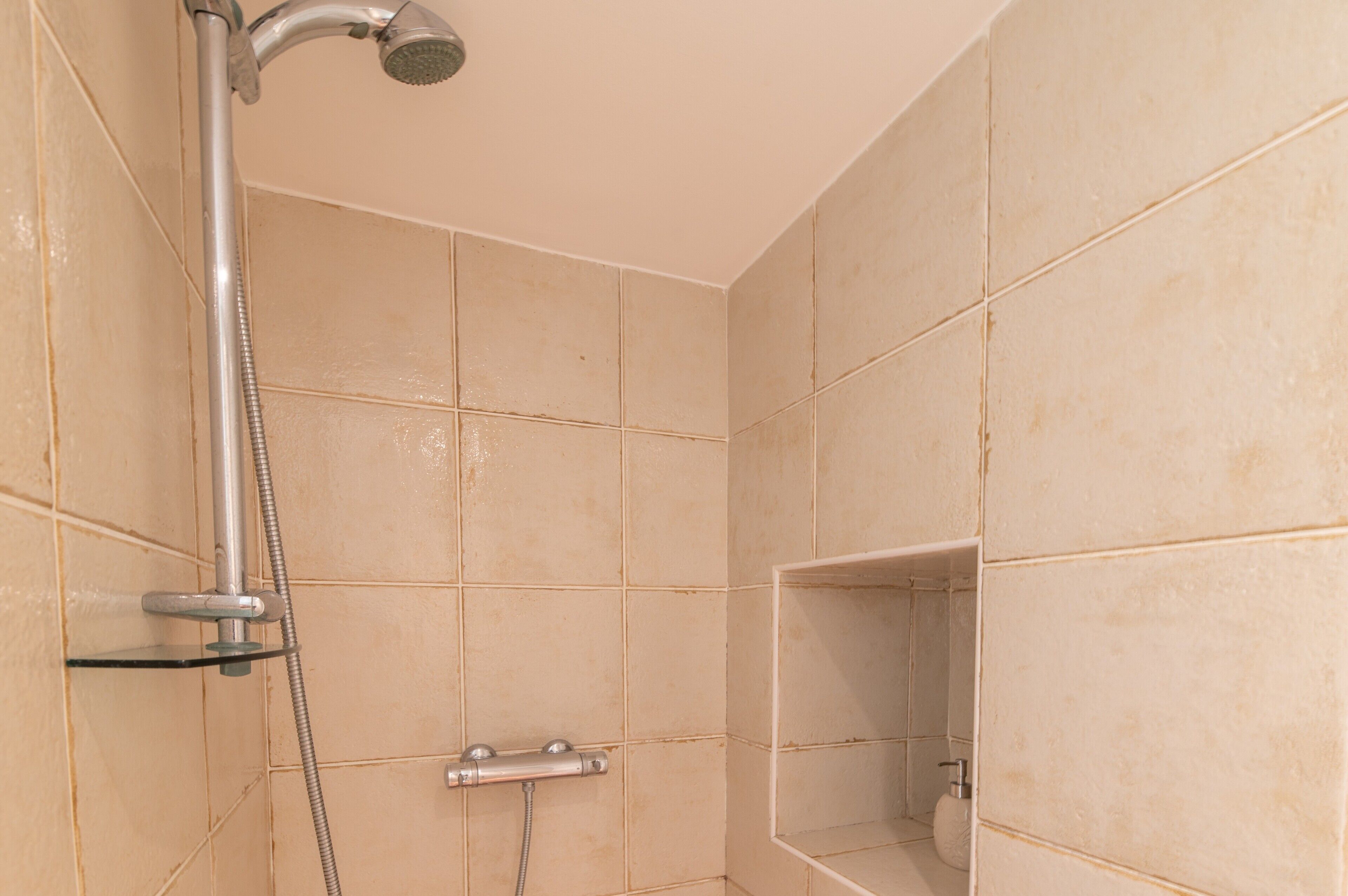 A shower with a handheld showerhead, a glass shelf, and a wall-mounted faucet.