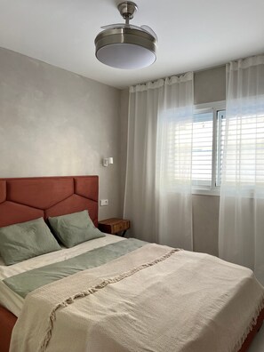 A bedroom with a bed, a headboard, a bedside table, a nightstand, a window with curtains, and a ceiling fan.