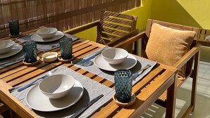 Una mesa de comedor de madera dispuesta con platos, fuentes y cubiertos, una silla de madera con cojín, y una valla de bambú al fondo.