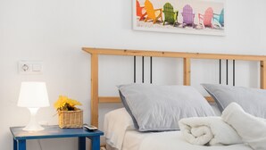 Une chambre avec une tête de lit en bois, une table de chevet avec une lampe et un panier, et un tableau représentant des chaises Adirondack aux couleurs vives accroché au mur.